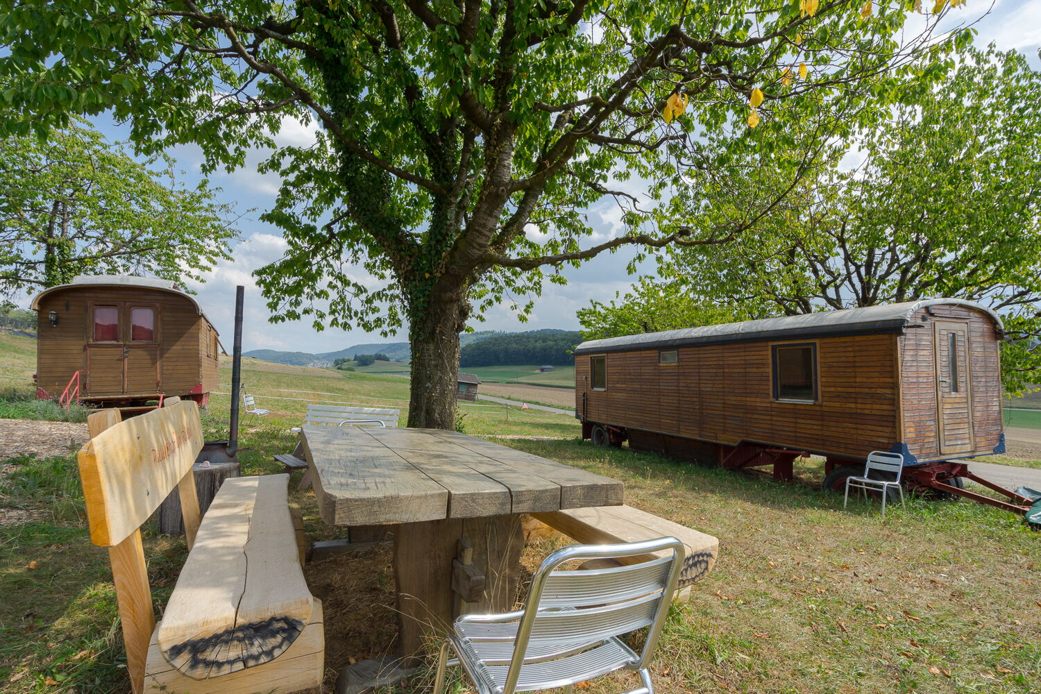 Zwei renovierte Zirkuswagen sowie Holztisch mit Bänken unter Bäumen.