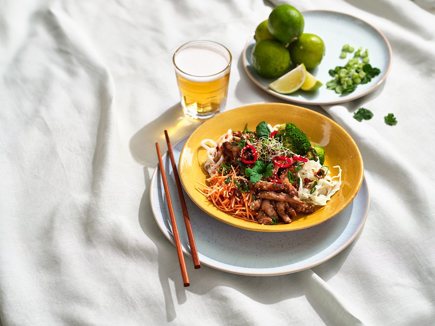 Plat de légumes et d'émincés joliment composé en couleurs dans un bol jaune. A côté, des baguettes et, en arrière-plan, une assiette de citrons verts.